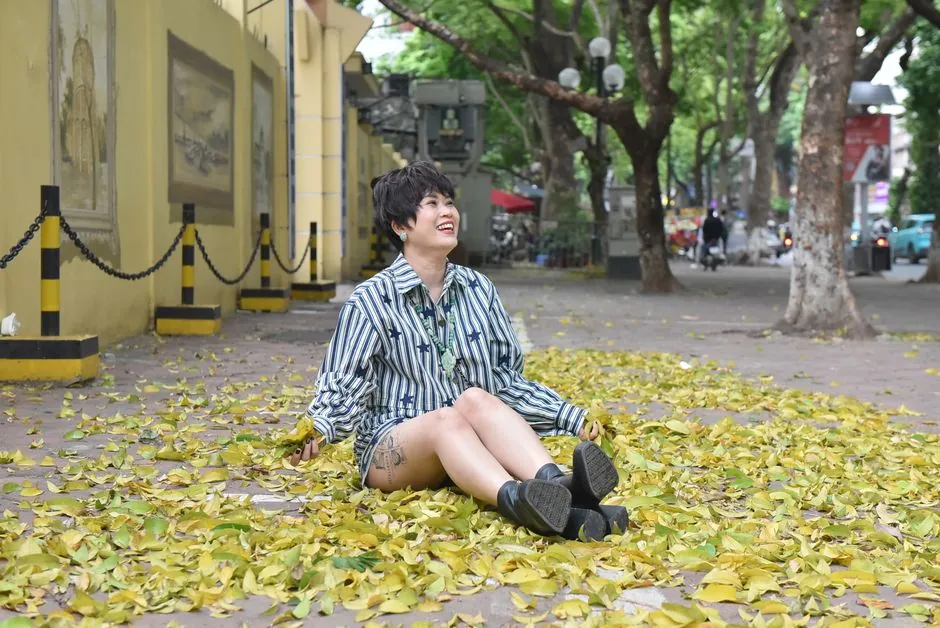 Smiling woman in striped shirt sitting on a leafy sidewalk in an urban setting.